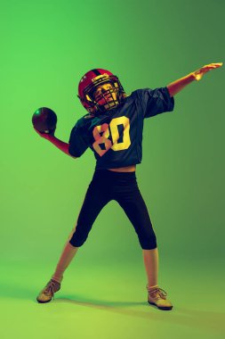 Sağlam atış. Spor üniformalı ve ekipmanlı Amerikan futbolu oynayan küçük bir çocuk neon ışıkta yeşil arka planda izole edilmiş. Spor, reklam, hareket, başarı kavramı.
