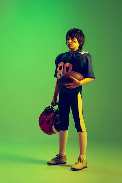 スポーツ少年の完全な長さの肖像画 ネオンの光の中で緑の背景に隔離された青のスポーツの制服や保護具でアメリカのサッカー選手 スポーツ 成果の概念 — ストック写真