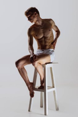 Üzücü ve ciddi görünüyor. Gri arka planda izole edilmiş yüksek sandalyede oturan beyaz şortlu, üstsüz, sportif bir genç. Sağlık, refah, insanın doğal güzelliği. Sanat, moda, çeşitlilik