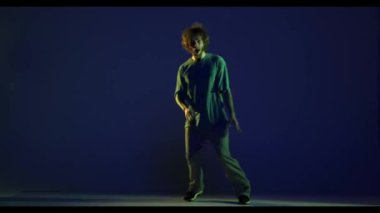 Duygular. Şık bir genç adam, spor kıyafeti giymiş hip-hop dansçısı koyu mavi arka planda izole bir şekilde dans ediyor. Modern dans sanatı, moda, gençlik, reklam, tarz konsepti. 4K, video