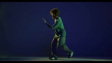 Şık bir genç adam, spor kıyafeti giymiş hip-hop dansçısı koyu mavi arka planda izole bir şekilde dans ediyor. Modern dans sanatı, moda, gençlik, reklam, tarz konsepti. 4K, video