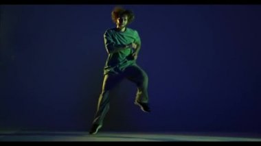 Atlıyorum. Şık bir genç adam, spor kıyafeti giymiş hip-hop dansçısı koyu mavi arka planda izole bir şekilde dans ediyor. Modern dans sanatı, moda, gençlik, reklam, tarz konsepti. 4K, video