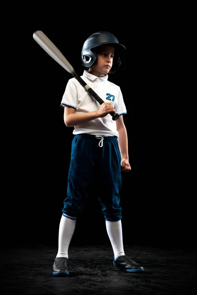 Full-length portrait of kid, beginner baseball player in sports uniform posing with baseball bat isolated on dark background. Concept of sport, achievements, competition. Future champion