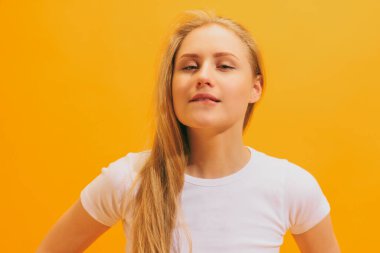 - Yakın çekim. Uzun sarı saçlı, beyaz tişörtlü, sarı arka planda yalnız poz veren duygusal genç kız. İnsan duyguları kavramı, yüz ifadesi, gençlik, arzu, reklam