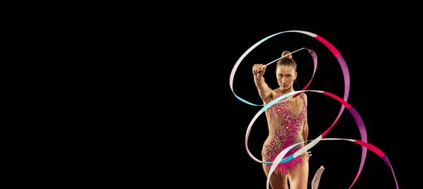 Solo performance. One young sportive girl, rhythmic gymnastics artist performing with ribbon isolated on dark background. Concept of sport, action, aspiration, education, active lifestyle