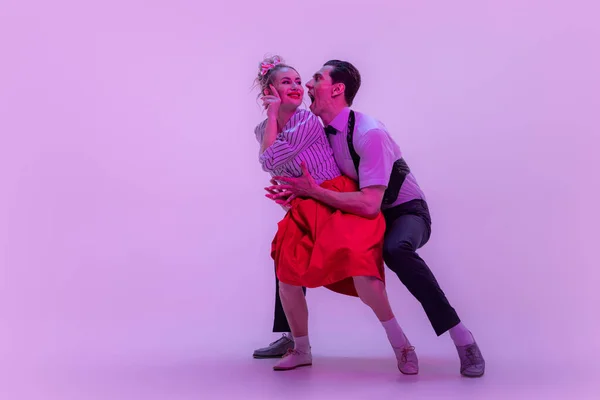 Social dancing. Studio shot of young man and woman in vintage retro style outfits dancing lindy hop isolated on lilac color background in neon light. Timeless traditions, 1960s fashion style and art