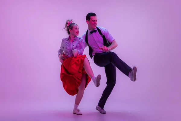Social dancing. Studio shot of young man and woman in vintage retro style outfits dancing lindy hop isolated on lilac color background in neon light. Timeless traditions, 1960s fashion style and art