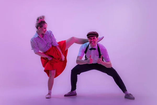 Social dancing. Studio shot of young man and woman in vintage retro style outfits dancing lindy hop isolated on lilac color background in neon light. Timeless traditions, 1960s fashion style and art