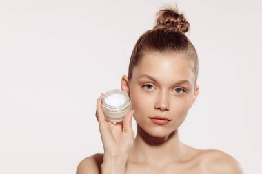 Makyajsız genç kızın yarım boy portresi beyaz stüdyo arka planında izole edilmiş krema şişesi tutuyor. Doğal kozmetik tedavisi. Güzellik kavramı, cilt bakımı, kozmetoloji, kaplıca