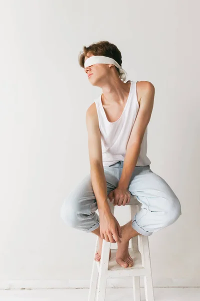 Sadness. Young man with blindfold sitting on chair isolated on white studio background. Concept of fashion, youth, emotions, facial expression, ad. Fashion male model in casual style clothes