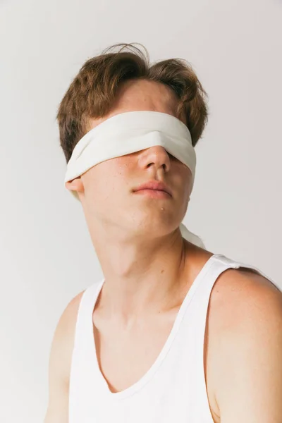 Closeup. Portrait of young man with blindfold sitting on chair isolated on white studio background. Concept of fashion, youth, emotions, facial expression, ad. Fashion model in casual style clothes