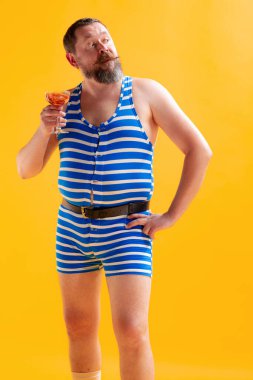 Mutlu ve eksantrik görünüyor. Komik denizci, eski çizgili mayo giyen şişman neşeli adam parlak sarı arka planda izole edilmiş poz veriyor. Tatil, yaz, komik mim duyguları konsepti.