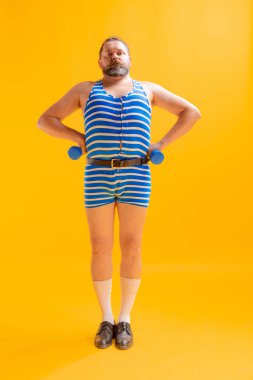 Merhamet. Retro çizgili mavi-beyaz mayo giyen yakışıklı şişman bir adam parlak sarı arka planda izole edilmiş spor egzersizleri yapıyor. Vücut pozitifliği, tatil, yaz, komik mim duyguları konsepti.