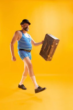 Yaz gezisi. Komik bir kumsala giden, retro çizgili mayo giyen ve parlak sarı arka planda izole edilmiş eski bir bavulu tutan şişman, heyecanlı bir adam. Tatil, duygular, reklam kavramı.