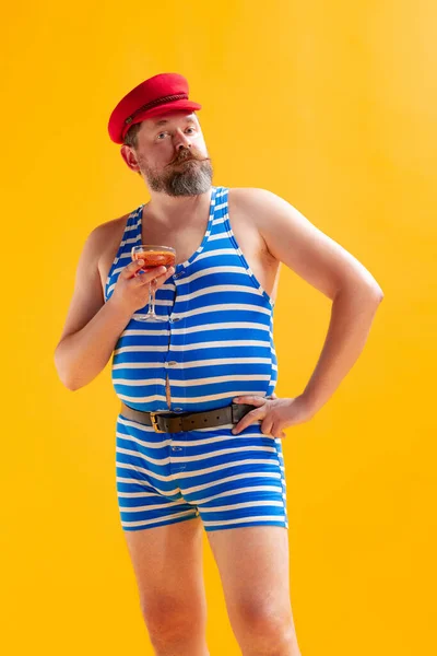 Portrait of funny eccentric seaman, fat cheerful man wearing retro striped swimsuit posing isolated on bright yellow background. Vacation, summer, funny meme emotions concept.