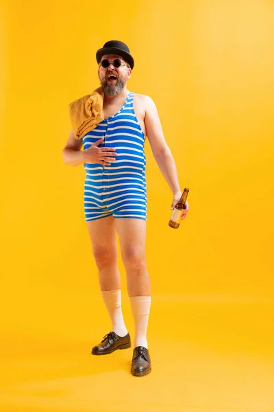 Laughing. Funny beachgoer, fat funny man wearing retro striped swimsuit and vintage bowler hat posing isolated on bright yellow background. Vacation, summer, funny meme emotions concept.