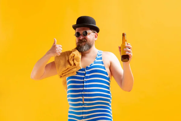 Tasting beer. Funny beachgoer, fat funny man wearing retro striped swimsuit and vintage bowler hat posing isolated on bright yellow background. Vacation, summer, funny meme emotions concept.