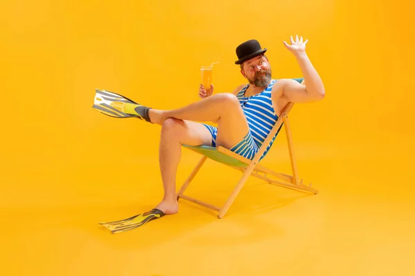 冷たいビールを味わう クールな面白いビーチガーのスタジオ撮影 ヴィンテージストライプの水着と黄色の背景に孤立サンベッドに座ってボウラー帽子の男 レジャー活動 — ストック写真