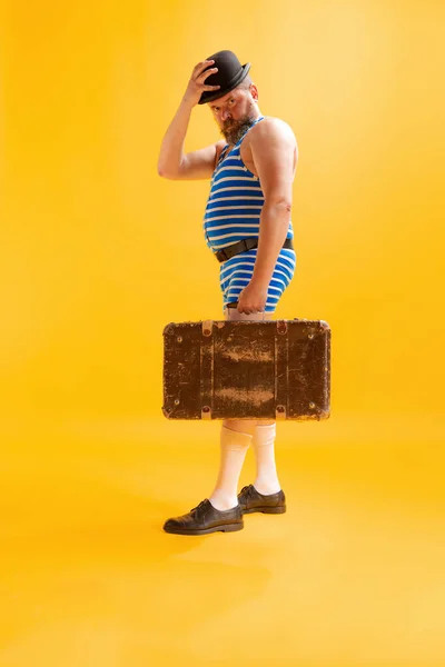 Summer trip. One funny beachgoer, fat excited man wearing retro striped swimsuit and vintage bowler hat holding old suitcase isolated on bright yellow background. Vacation, emotions, ad concept.