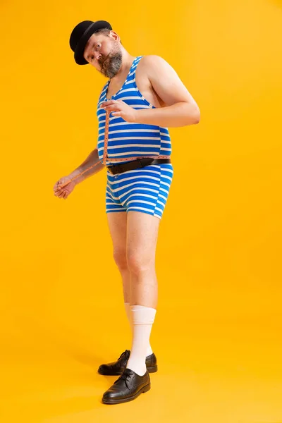 Stock image Measurement. Cute fat young man wearing retro striped swimsuit and vintage bowler hat measuring himself isolated on bright yellow background. Vacation, summer, healthy diet concept.