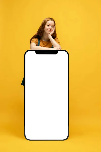 New opportunity. Young happy girl next to huge 3d model of smartphone with empty white screen isolated on yellow background, Fashion, new app or website, copy space for your ad, mockup