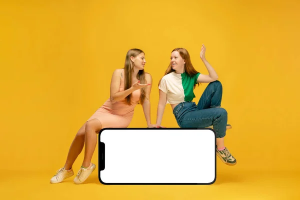 Advice to friend Two excited young girl sitting on huge 3d model of cellphone with blank white screen isolated on yellow background, Fashion, new app or website, copy space for ad, mockup or design