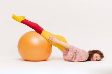 Top üzerinde alıştırma yapıyorum. Avangard tarzı giyinmiş, beyaz arka planda eğlenen oldukça komik bir genç kız. Fitness, canlı tarz, güzellik, nonoş, ucube, moda konsepti.