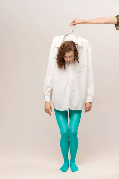 Marionette, puppet. Young girl in white shirt and blue tights controlled by the other hand isolated over white background. Vivid style, beauty, queer, freak, fashion concept.