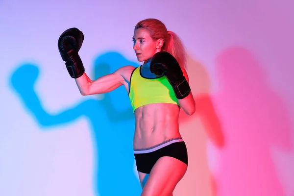 Right hook. Energetic young woman, female professional kickboxer in sports uniform and gloves practicing isolated on pink background in neon with shadow. Sport, competition, power concept.
