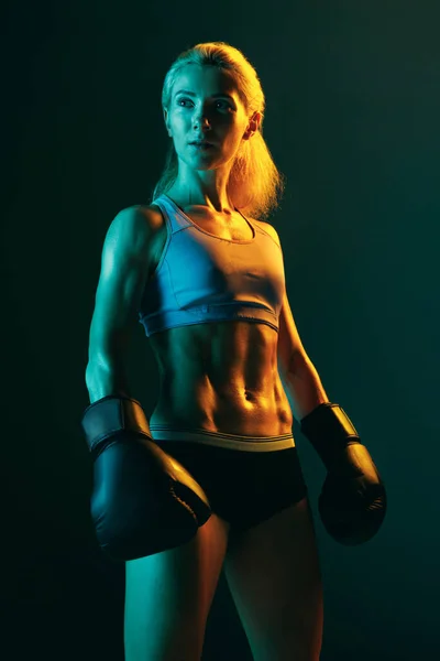 Beauty and sport. One young woman, female professional kickboxer in sports uniform and gloves training isolated on dark green background in neon. Competition, power concept.