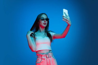 Selfie çekiyorum. Uzun, parlak siyah saçlı, mavi arka planda, neon ışıkta izole edilmiş duygusal bir kızın yarım boy çekimi. Güzellik, sanat, moda, gençlik, satış ve reklamlar kavramı.