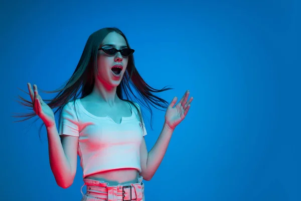 Wow emotions. Excited young girl with long glossy dark hair shouting isolated on blue background in neon light. Concept of beauty, art, fashion, youth, sales and ads.