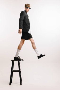 İlk adım, belirsizlik. Genç havalı bir adam, siyah şortlu ve ceketli beyaz arka planda izole bir şekilde yüksek sandalyede duran bir öğrenci. Sanat, güzellik, moda, gençlik kültürü, satış ve reklamlar