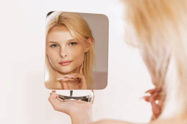 Close-up portrait of young age beautiful tow-head girl looks at her face in mirror. Beauty, spa, age-related changes, face lifting and skin care concept.