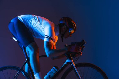 Closeup portrait of professional male cyclist in sports uniform, goggles and helmet on blue background in neon. Concept of active life, rest, travel, energy, sport. Copy space for ad. Moon light.