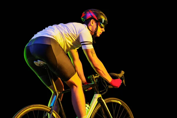 Sportsman. Portrait of young male cyclist on bicycle in cycling shorts and protective helmet isolated on dark background in neon light. Concept of active life, rest, travel, energy, sport.