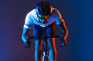 Closeup portrait of professional male cyclist in sports uniform, goggles and helmet on blue background in neon. Concept of active life, rest, travel, energy, sport. Copy space for ad. Front view