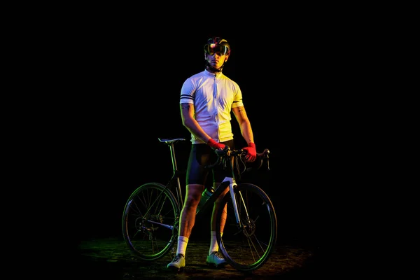 Biker. Full-length portrait of young male cyclist on bicycle in cycling shorts and protective helmet isolated on dark background in neon light. Concept of active life, rest, travel, energy, sport.