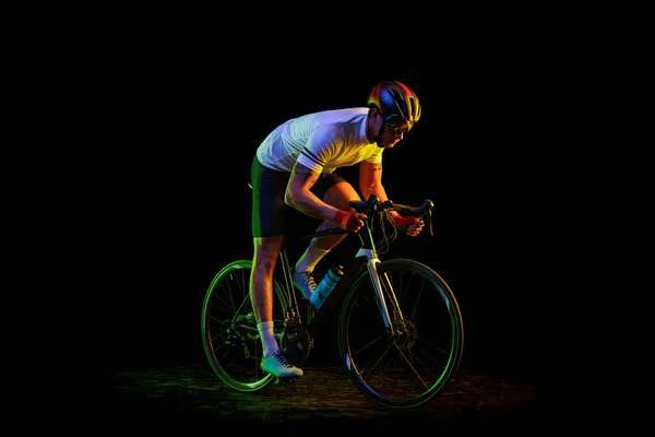 Sportsman. Portrait of young male cyclist on bicycle in cycling shorts and protective helmet isolated on dark background in neon light. Concept of active life, rest, travel, energy, sport.