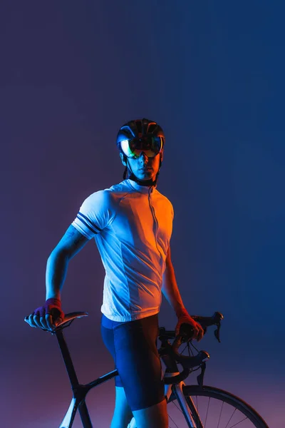 Rest time. One male cyclist riding bicycle wearing cycling shorts and protective helmet isolated on dark blue background in neon. Concept of active life, rest, travel, energy, sport. Copy space for ad