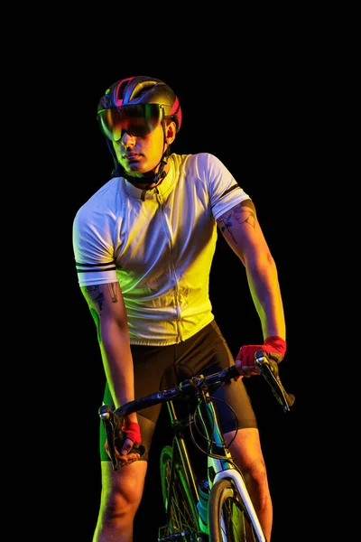 Sportsman. Portrait of young male cyclist on bicycle in cycling shorts and protective helmet isolated on dark background in neon light. Concept of active life, rest, travel, energy, sport.