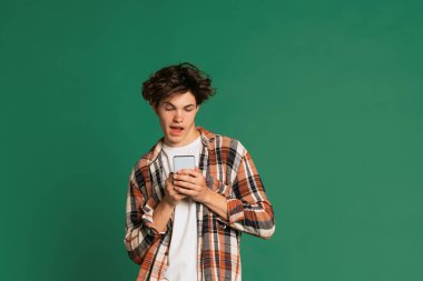Cep telefonuyla. Cinsiyet z. Genç bir çocuk, yeşil stüdyo arka planında izole edilmiş günlük kıyafetlerle poz veriyor. Okula dönmek, duygular, gençlik, moda konsepti. İlan için alanı kopyala