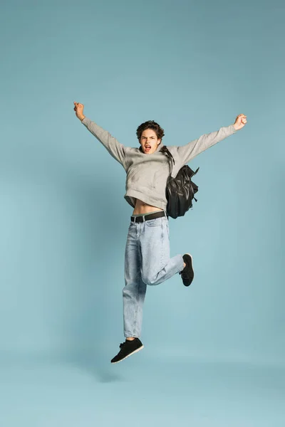 Excitement. Happy student, teen in jeans and grey hoodie jumping isolated over blue background. Concept of emotions, facial expression, youth. Back to school. Model with leather backpack
