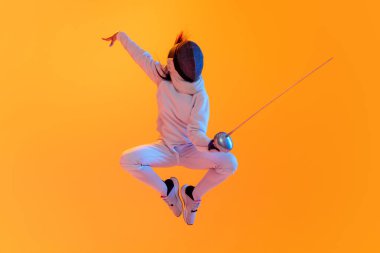 Jumping. Studio shot of professional fencer in white fencing costume and mask in action, motion isolated on orange color background. Concept of sport, youth, activity, skills, achievements, ad.