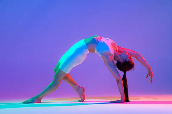 Stretching. Studio shot of young flexible girl in fitness sport uniform practicing isolated on gradient pink-purple background in neon. Modern sport, action, fitness, yoga, motion, youth concept.