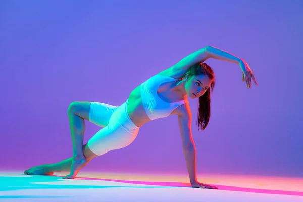 Studio shot of young flexible girl in fitness sport uniform practicing isolated on gradient pink-purple background in neon light. Modern sport, action, fitness, yoga, motion, youth concept.