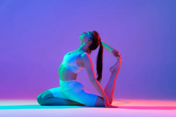 Yoga pose. Studio shot of young flexible girl in fitness sport uniform practicing isolated on gradient pink-purple background in neon light. Modern sport, action, fitness, yoga, motion, youth concept.