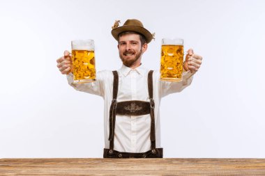 Young expressive man in hat, wearing fest traditional Bavarian or German costume with big beer glass isolated on white background. Alcohol, traditions, holidays, Oktoberfest concept. Copy space for ad