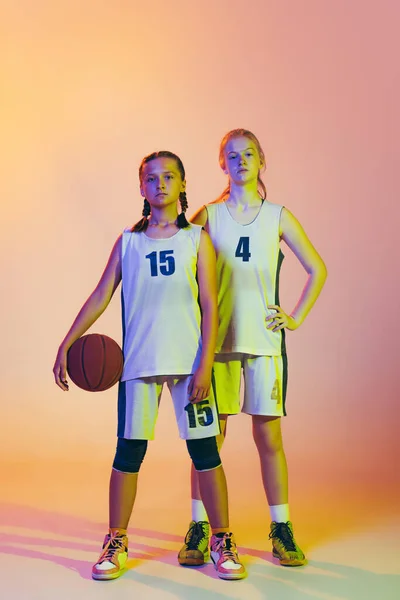 First team. Two sportive girls, teens, basketball players standing together with basketball ball isolated on pink background in neon light. Concept of sport, team, enegry, skills. Juniors.