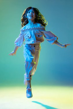 Running. Beautiful fashionable little girl, kid posing isolated over cyan color background in white neon light. Concept of children emotions, fashion, beauty, back to school and ad